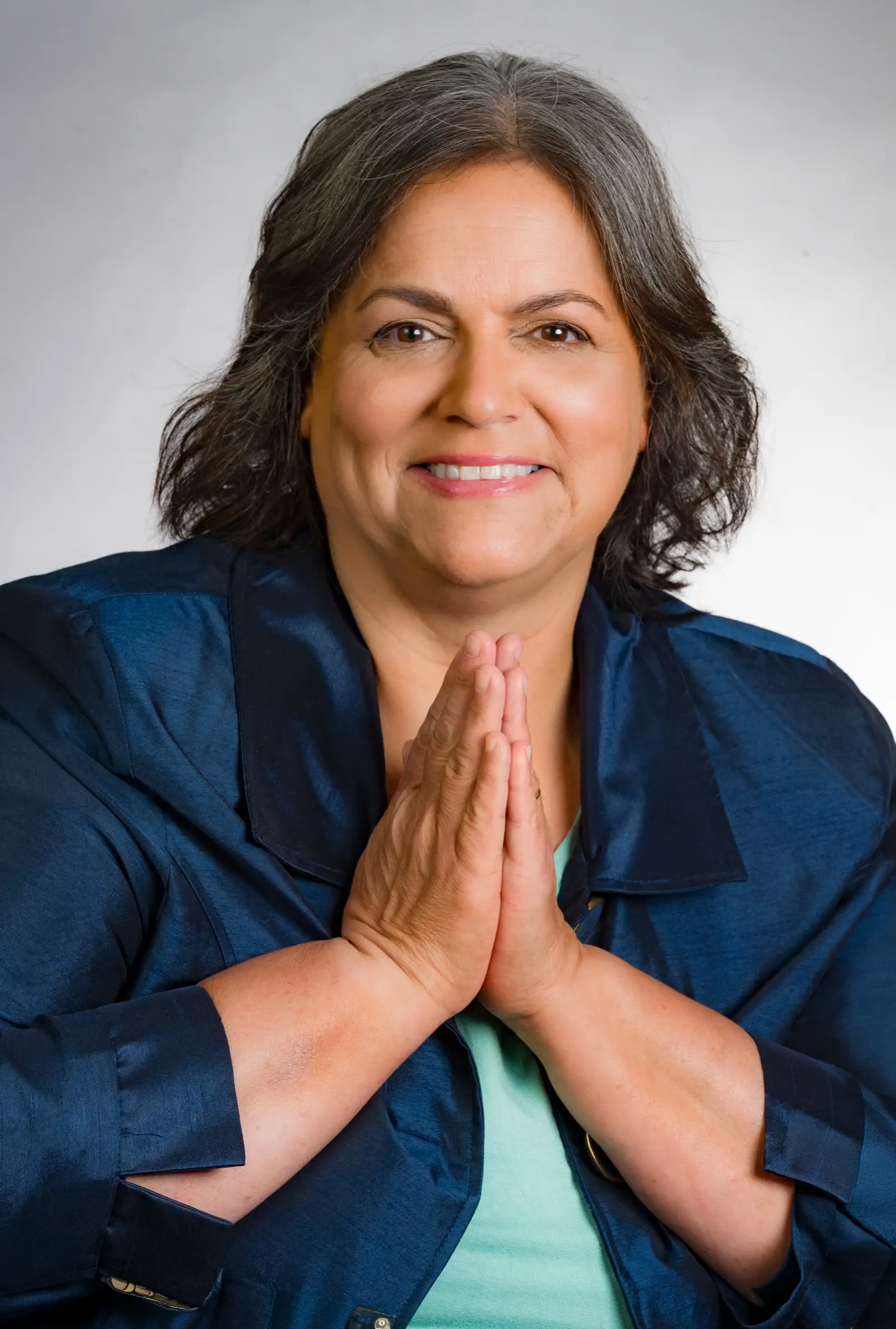 Studio portrait of Esmeralda Reynolds smiling with palms together, wearing a navy jacket over a mint top